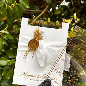 May include: Wedding invitation with gold text on a white card, featuring the names "Mohamed & Memuna". The card is adorned with a white ribbon, dried flowers, and a gold seal. A clear overlay displays additional details.