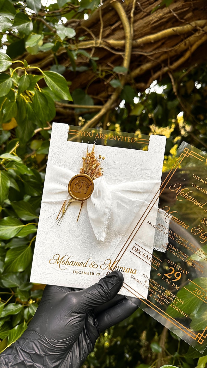 May include: Wedding invitation with gold text on a white card, featuring the names "Mohamed & Memuna". The card is adorned with a white ribbon, dried flowers, and a gold seal. A clear overlay displays additional details.
