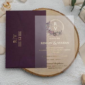 Könnte beinhalten: Eine Hochzeitseinladungssuite mit einem weinroten Umschlag und goldenem Text sowie einer transparenten Folie. Die Einladung enthält die Initialen "R" und "V", das Datum und die Namen. Goldene Schere und Band sind ebenfalls vorhanden.