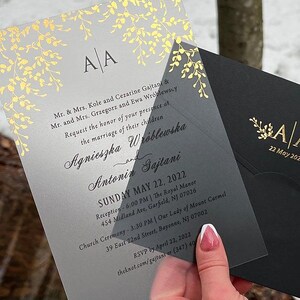 May include: A clear wedding invitation with gold foil leaf accents and the initials "A A". The invitation includes the names of the couple, the date, and the location of the ceremony and reception. A black envelope is visible.