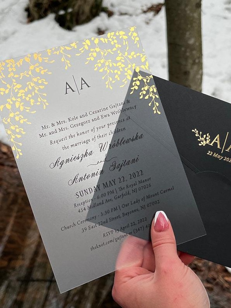 May include: A clear wedding invitation with gold foil leaf accents and the initials "A A". The invitation includes the names of the couple, the date, and the location of the ceremony and reception. A black envelope is visible.