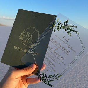 Könnte beinhalten: Zwei Hochzeitseinladungen werden gezeigt, eine dunkelgrüne und eine transparente. Die dunkelgrüne Einladung trägt den Text "ROSA & ANUP" und "08.19.2023". Die transparente Einladung trägt den Text "ROSA TAMANG & ANUP GAZMERE". Beide haben florale Designs.