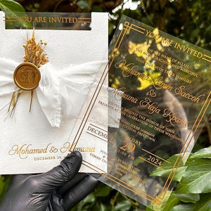 May include: Elegant wedding invitations with gold lettering on clear acrylic and white cardstock. The invitations feature the text "You are Invited" and include details for the wedding ceremony and reception. A white ribbon and dried flowers adorn one invitation.