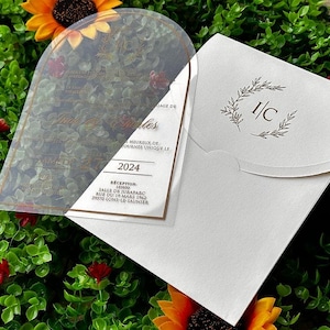 May include: A clear acrylic wedding invitation with gold text and a white envelope. The invitation includes the year 2024 and the text "Heureux de". The envelope has a floral design and the initials "I/C".