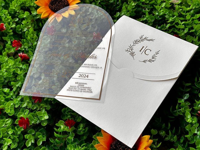 May include: A clear acrylic wedding invitation with gold text and a white envelope. The invitation includes the year 2024 and the text "Heureux de". The envelope has a floral design and the initials "I/C".