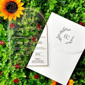 May include: A clear acrylic wedding invitation with gold accents and script. The invitation includes the names "Inas & Charles" and the date "Saturday 27 2024". A white envelope with a matching design is also present.