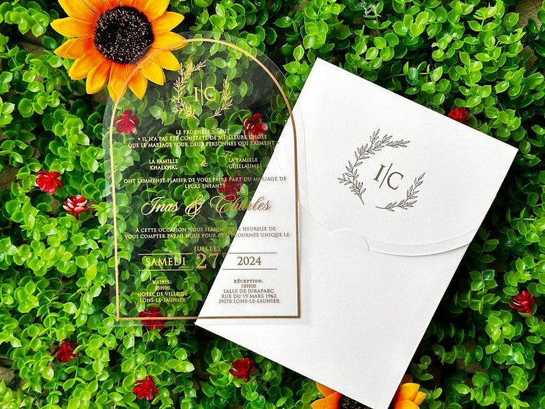 May include: A clear acrylic wedding invitation with gold accents and script. The invitation includes the names "Inas & Charles" and the date "Saturday 27 2024". A white envelope with a matching design is also present.