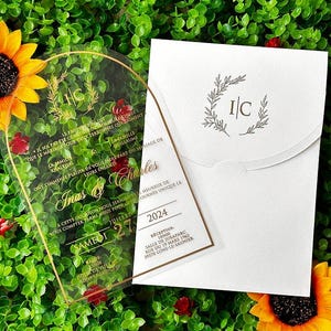 May include: A clear acrylic wedding invitation with gold lettering and a gold border, featuring the names "Inas & Charles" and event details. The invitation is paired with a white envelope, and the scene is set against a backdrop of greenery and sunflowers.