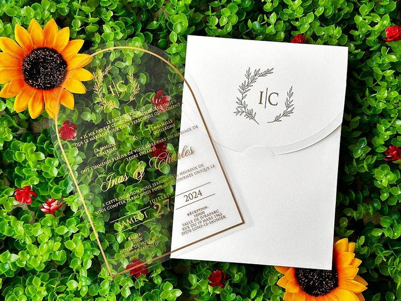 May include: A clear acrylic wedding invitation with gold lettering and a gold border, featuring the names "Inas & Charles" and event details. The invitation is paired with a white envelope, and the scene is set against a backdrop of greenery and sunflowers.