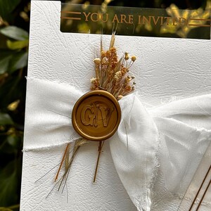 May include: A white invitation card with a gold wax seal and a white ribbon. The seal has the initials "GIV". The card is decorated with dried flowers and the text "YOU ARE INVITED". The names "Mohamed & Memuna" and the date "DECEMBER 29, 20" are also visible.