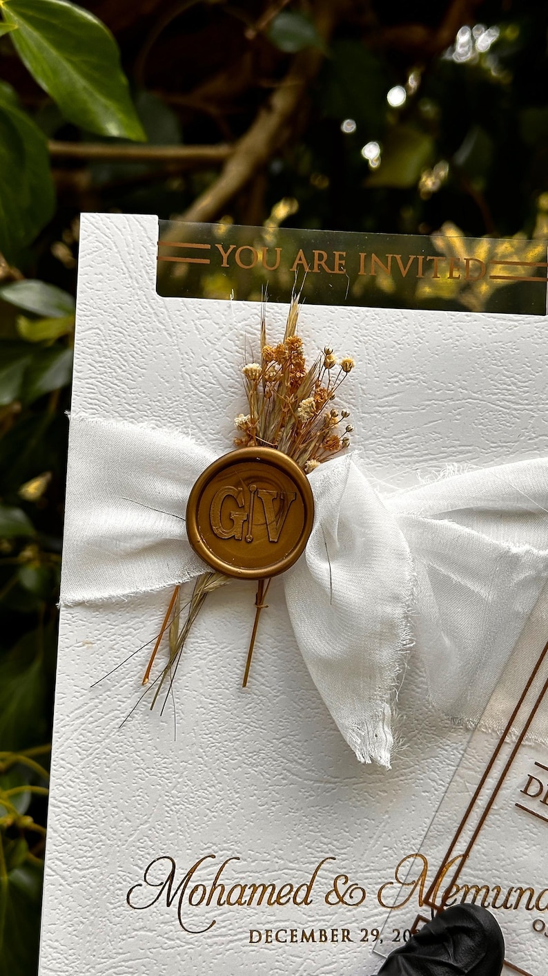 May include: A white invitation card with a gold wax seal and a white ribbon. The seal has the initials "GIV". The card is decorated with dried flowers and the text "YOU ARE INVITED". The names "Mohamed & Memuna" and the date "DECEMBER 29, 20" are also visible.