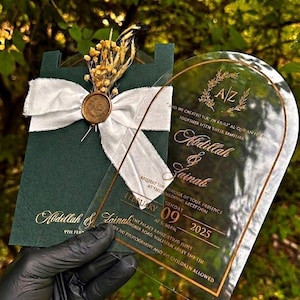 Puede incluir: Elegante invitaci&oacute;n de boda con una tarjeta verde oscuro con letras doradas, una cinta blanca y un sello dorado. Una tarjeta acr&iacute;lica transparente con texto dorado est&aacute; superpuesta. La invitaci&oacute;n incluye los nombres Abdillah & Zainab y la fecha 09 de febrero de 2025.