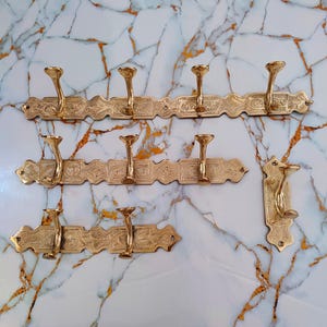 May include: A collection of brass-coloured wall-mounted coat racks with multiple hooks. The racks feature an ornate, engraved design and are displayed on a marble-patterned surface. The hooks are curved and flared.