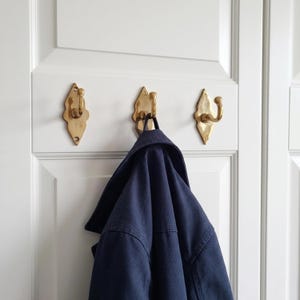 May include: Three ornate, brass-colored wall hooks mounted on a white door. A navy blue jacket hangs from the center hook. The hooks have a decorative, leaf-like shape, adding a touch of elegance to the entryway.