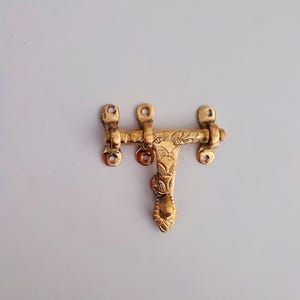 May include: A decorative brass door latch. The latch features an engraved floral pattern and has a polished, golden finish. It consists of a horizontal bar with mounting holes and a vertical piece with a decorative handle.