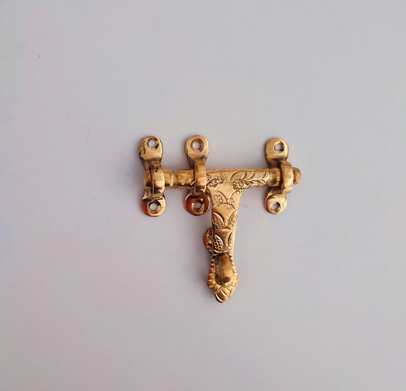 May include: A decorative brass door latch. The latch features an engraved floral pattern and has a polished, golden finish. It consists of a horizontal bar with mounting holes and a vertical piece with a decorative handle.
