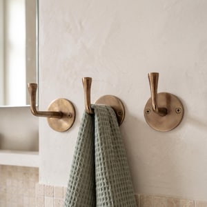 May include: Three brass towel hooks mounted on a light-colored wall. One hook is holding a green waffle-weave towel. The hooks have a circular base and a curved, upward-facing hook. The wall has a textured finish, and a mirror is partially visible.