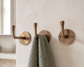 May include: Three brass towel hooks mounted on a light-colored wall. One hook is holding a green waffle-weave towel. The hooks have a circular base and a curved, upward-facing hook. The wall has a textured finish, and a mirror is partially visible.
