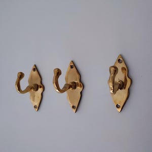 May include: Three brass wall hooks with a diamond-shaped backplate and curved hook. The hooks are a warm gold color and are mounted on a light gray wall. Each hook has a decorative, scalloped edge.