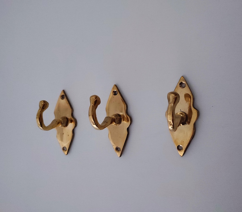 May include: Three brass wall hooks with a diamond-shaped backplate and curved hook. The hooks are a warm gold color and are mounted on a light gray wall. Each hook has a decorative, scalloped edge.