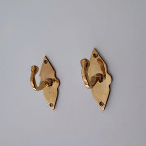 May include: Two brass wall hooks with a diamond-shaped backplate and a curved hook. The hooks are a shiny gold color and have a decorative edge. Each hook has two screw holes at the bottom and one at the top for mounting.