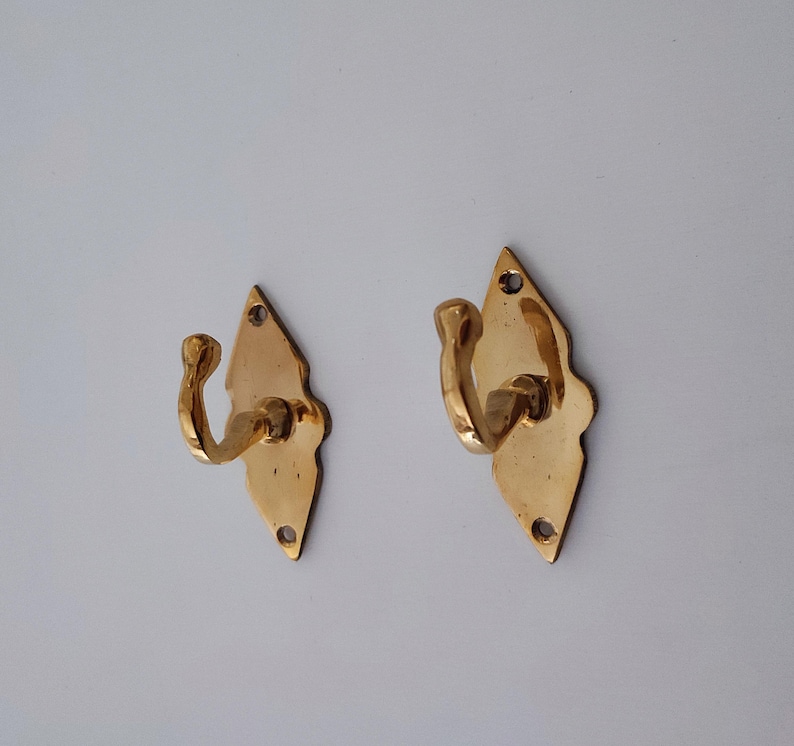 May include: Two brass wall hooks with a diamond-shaped backplate and a curved hook. The hooks are a shiny gold color and have a decorative edge. Each hook has two screw holes at the bottom and one at the top for mounting.