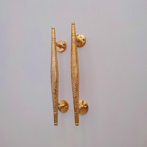 Peut inclure: Deux poignées de porte dorées avec des motifs floraux complexes. Chaque poignée est allongée, avec des extrémités arrondies et une base circulaire. Les poignées sont symétriques et semblent être en métal.