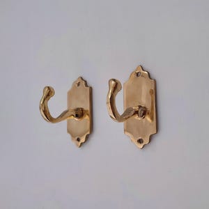 May include: Two brass-colored wall hooks. Each hook has a curved design and is mounted on a rectangular backplate with decorative edges. The hooks are made of metal and are designed for hanging items.