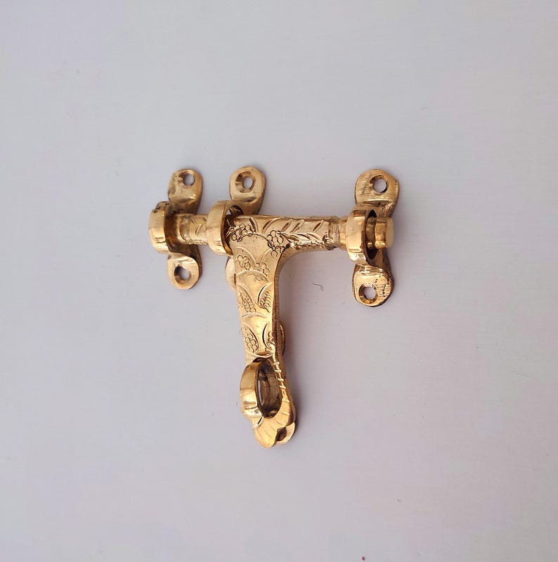 May include: A gold-coloured brass door latch with an intricate floral pattern. The latch has a sliding bolt mechanism. The decorative hardware is mounted on a white surface.