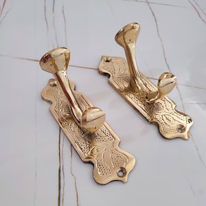 May include: Two brass-colored wall hooks with a decorative floral design. Each hook has a curved arm and a rounded top. The rectangular backplates are also adorned with floral patterns and have a hole for mounting. The hooks are made of metal.