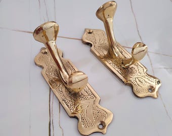 May include: Two brass-coloured wall hooks with a decorative floral design. Each hook has a curved arm and a rounded top. The rectangular backplates are also adorned with floral patterns and have a hole for mounting. The hooks are made of metal.