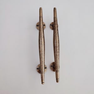 May include: Two bronze-colored door handles with intricate floral patterns. Each handle has a long, curved design with decorative detailing and is mounted on a circular base. The handles are symmetrical and appear to be made of metal.