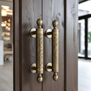 May include: A pair of antique brass door handles with detailed engraved designs. The handles are cylindrical, featuring decorative finials and circular backplates. The door is made of dark stained wood.