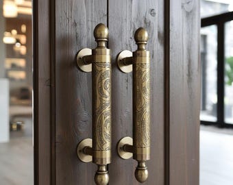 Pode incluir: Duas maçanetas de porta antigas em latão com padrões gravados intrincados. As maçanetas são cilíndricas com pináculos decorativos e placas traseiras circulares. A porta é de madeira escura manchada.