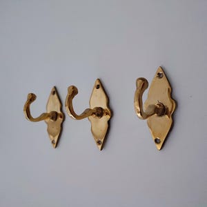 May include: Three brass wall hooks with a decorative diamond-shaped backplate. The hooks have a curved design and are mounted on a white wall. The hooks are made of a shiny, gold-colored metal.
