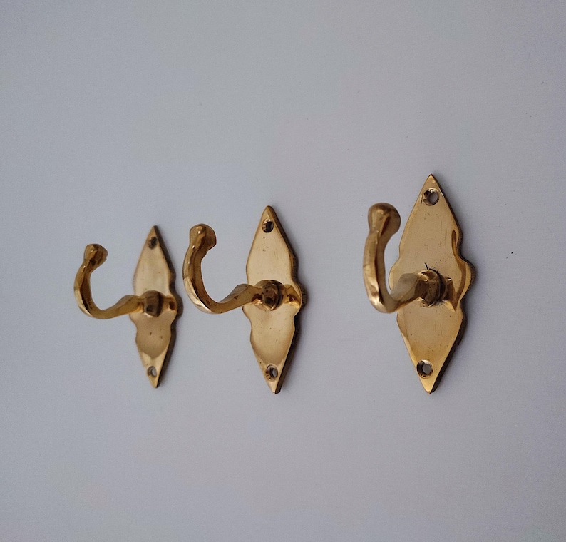 May include: Three brass wall hooks with a decorative diamond-shaped backplate. The hooks have a curved design and are mounted on a white wall. The hooks are made of a shiny, gold-colored metal.