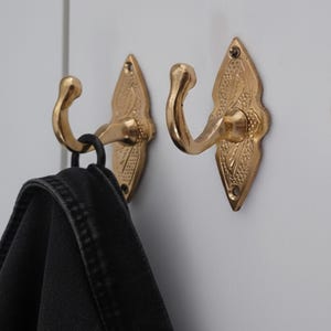 May include: Two ornate, gold-coloured metal wall hooks with a decorative leaf design. One hook holds a black garment. The hooks are mounted on a white surface, providing a classic and functional storage solution.