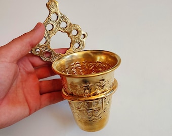 May include: A gold-colored metal cup with intricate embossed designs and a decorative handle. The cup has a wide rim and a detailed pattern around the body. The handle features a geometric openwork design. The cup is held against a plain white background.