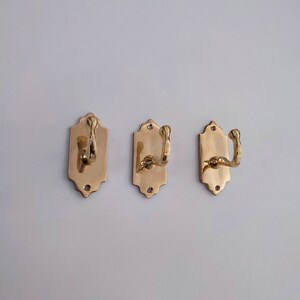 May include: Three brass wall hooks. Each hook is mounted on a rectangular backplate with a decorative top edge. The hooks have a curved design. The brass finish gives them a vintage look.