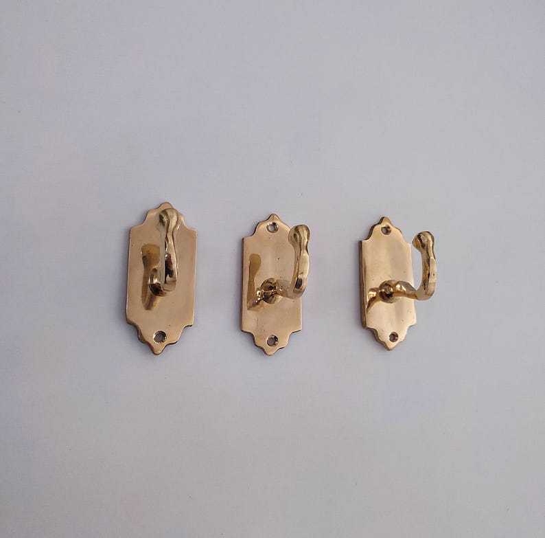 May include: Three brass wall hooks. Each hook is mounted on a rectangular backplate with a decorative top edge. The hooks have a curved design. The brass finish gives them a vintage look.
