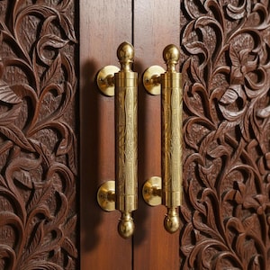 Puede incluir: Primer plano de un armario de madera con dos tiradores de latón. Los tiradores son cilíndricos con grabados decorativos y extremos redondeados. Las puertas del armario son de color marrón oscuro con diseños florales tallados.