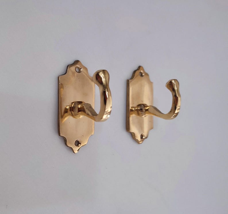 May include: Two brass-colored wall hooks. Each hook has a decorative backplate with a curved hook extending outward. The hooks are mounted on a white wall. The hooks are made of a shiny, metallic material.