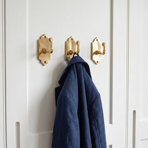 Puede incluir: Tres perchas de pared de color latón con un diseño decorativo están montadas en una puerta blanca. Una chaqueta azul marino cuelga de una de las perchas. Las perchas tienen un diseño curvo.