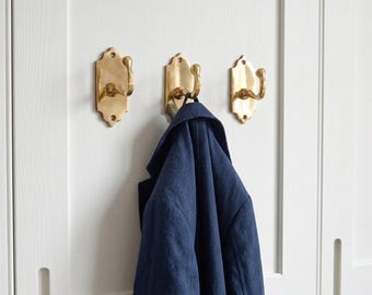 May include: Three brass-colored wall hooks with a decorative design are mounted on a white door. A navy blue jacket hangs from one of the hooks. The hooks have a curved design.