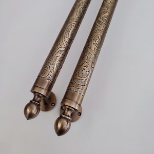 Könnte beinhalten: Zwei bronzefarbene Gardinenstangen mit kunstvollen, wirbelnden Mustern. Jede Stange hat eine dekorative Endkappe und Montagematerial. Die Stangen sind für Fensterdekorationen konzipiert und verleihen jedem Raum einen Hauch von Eleganz.
