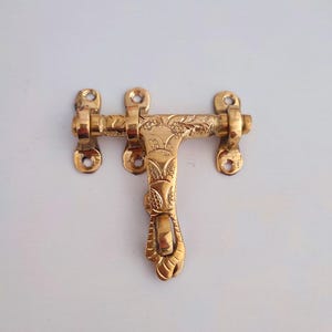 May include: A brass door latch with floral and leaf engravings. The latch has a rectangular bar with three screw holes on each end and a decorative handle. The metal has a polished, golden finish.