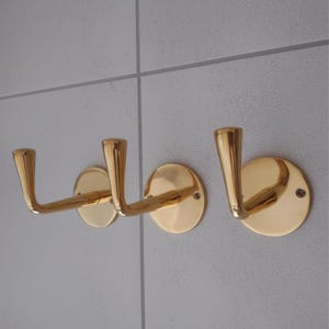 Puede incluir: Tres perchas de pared doradas. Cada gancho tiene un diseño curvo y está montado sobre una base redonda. Los ganchos están hechos de un material metálico brillante y están fijados a una pared gris claro.