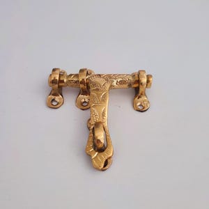 May include: A golden-coloured, ornate door latch. The latch features a T-shaped design with decorative floral patterns and a curved handle. The metal hardware has mounting holes for installation and is likely made of brass.
