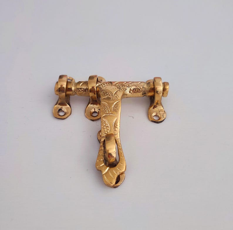 May include: A golden-coloured, ornate door latch. The latch features a T-shaped design with decorative floral patterns and a curved handle. The metal hardware has mounting holes for installation and is likely made of brass.