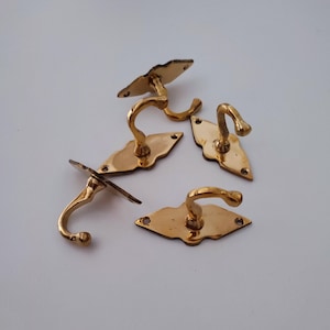 May include: A collection of five antique brass hooks. Each hook has a curved design and is mounted on a decorative backplate. The hooks are in various shapes, including diamond and oval, and are ideal for hanging items.